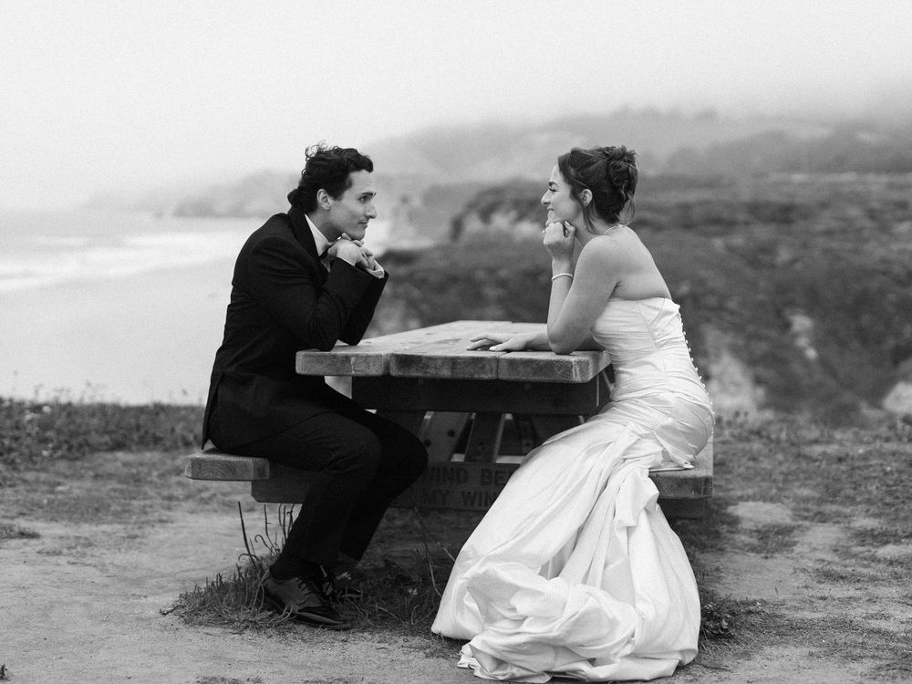 A black and white photo of newlyweds sitting at a rustic table with dramatic coastal views.