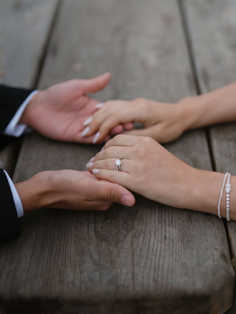 Close-up of hands with wedding rings and bracelets touching tenderly on wooden table surface.