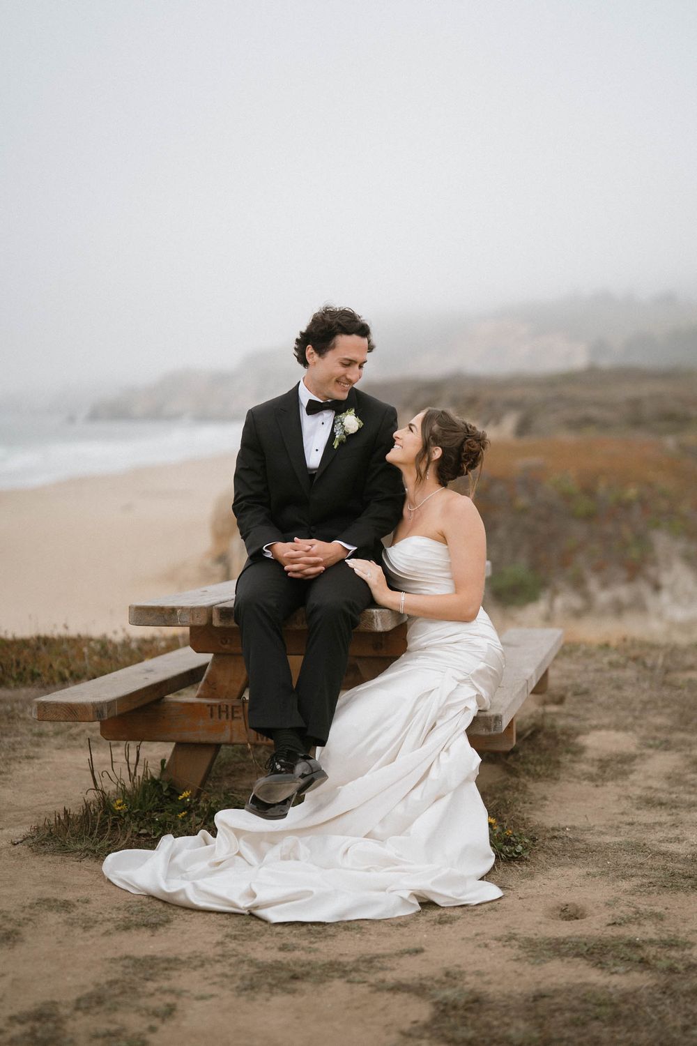 A couple in wedding attire sits together on a rustic bench along a misty coastal landscape.