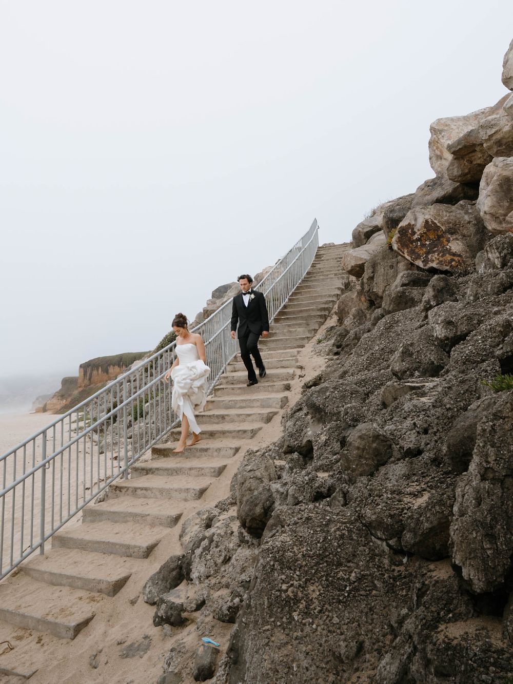 A couple in wedding attire walks up stone steps along a rocky coastal cliff with a misty ocean backdrop.