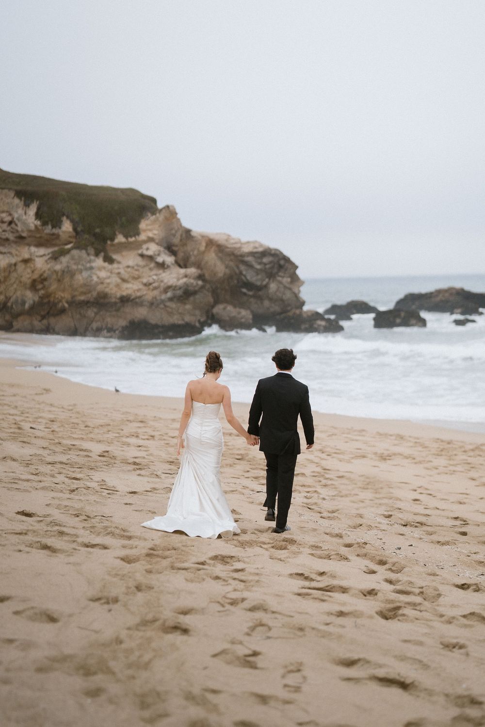 A couple in wedding attire walks along a dramatic coastal beach with rocky cliffs and crashing waves in the background.