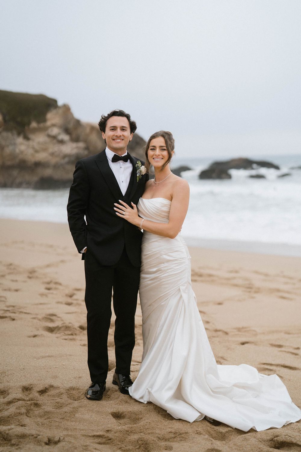A couple poses for their wedding photos on a scenic beach with cliffs and ocean waves in the background.