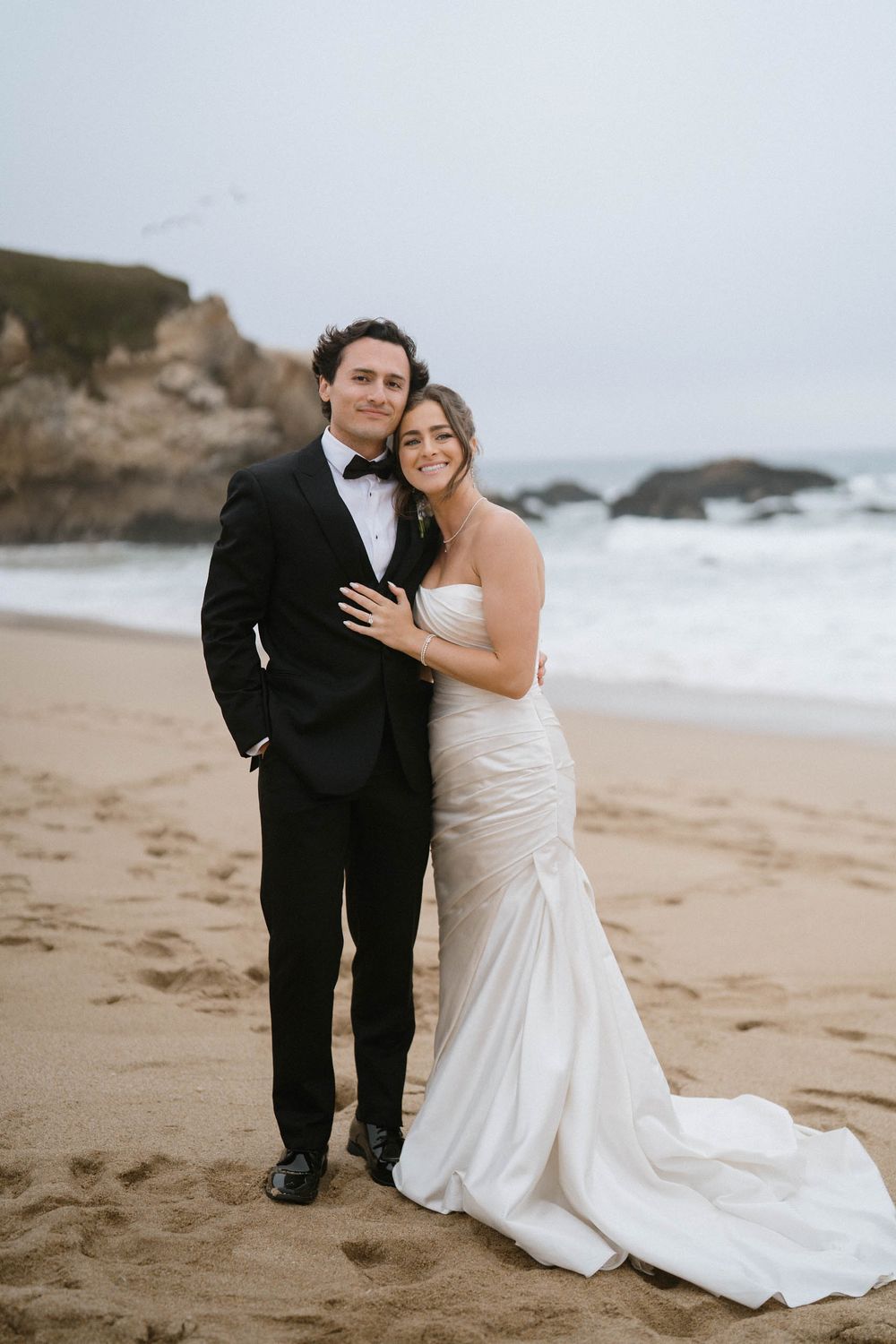 A romantic wedding couple embraces on a cloudy beach with rocky cliffs in the background.