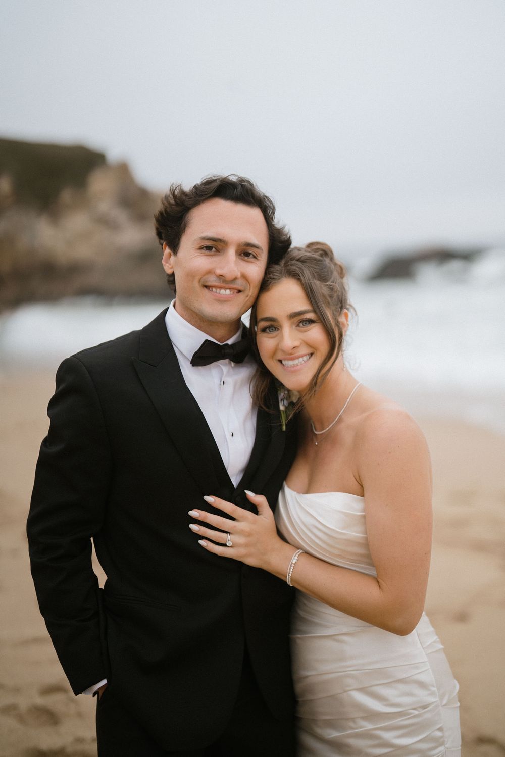A couple in formal wedding attire poses together on a beach during their romantic coastal photo session.