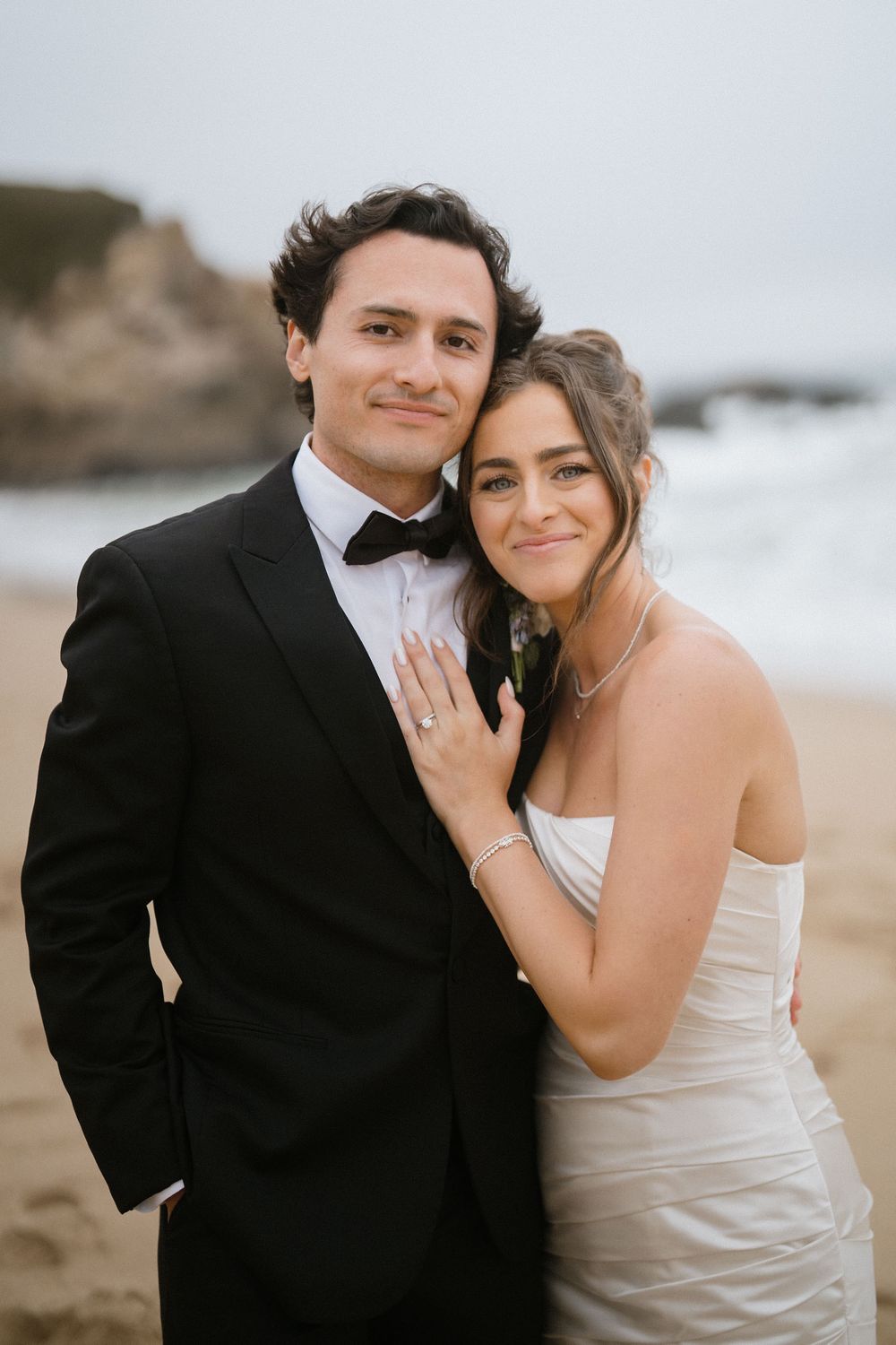 A romantic couple in wedding attire embraces on a beach with waves crashing in the background.