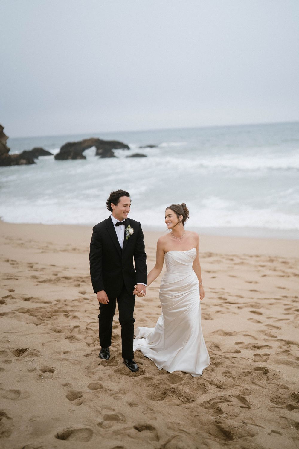 A couple walks together on a misty beach with crashing waves and rocky outcrops in the background.
