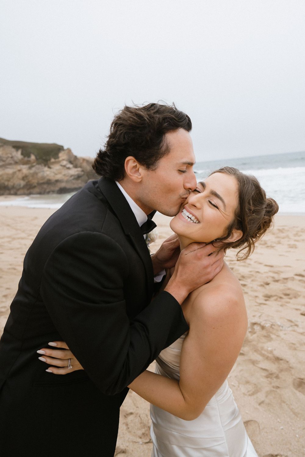 A couple shares intimate kisses during their beach wedding photoshoot with ocean waves behind them.