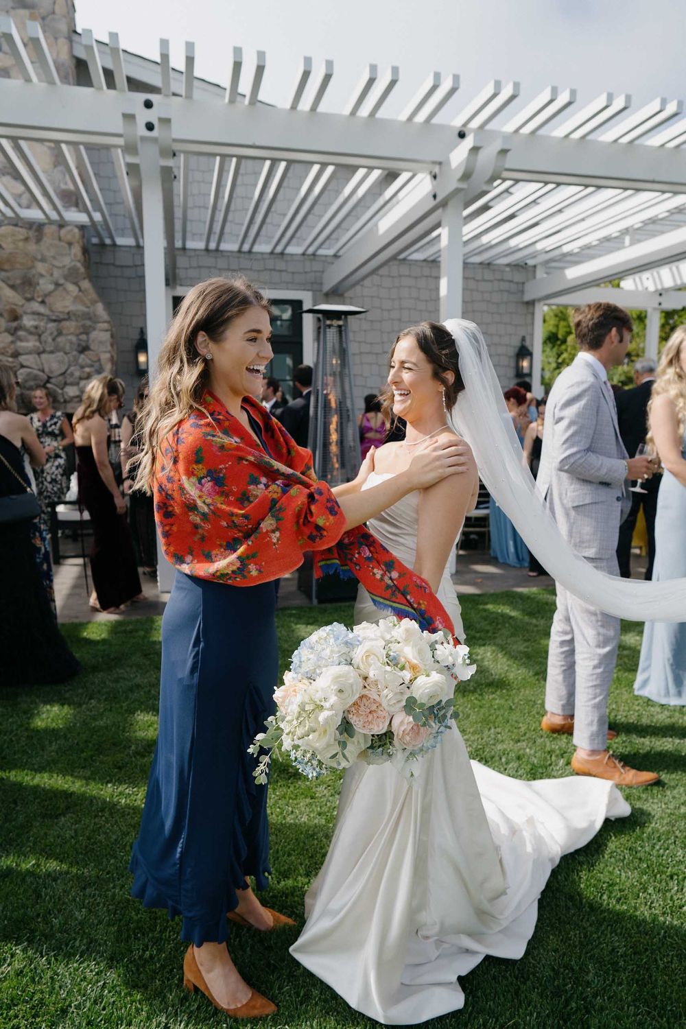 Wedding guests embrace and celebrate under a white pergola during an outdoor reception on a sunny day.