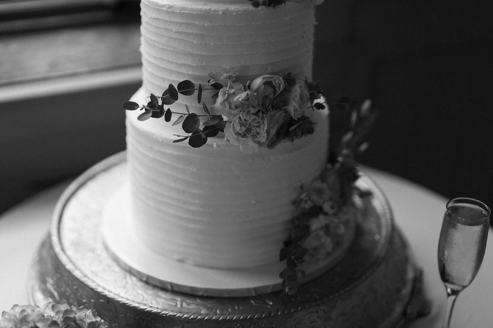 A black and white photograph of an elegant two-tiered wedding cake decorated with flowers and sitting next to a champagne glass.