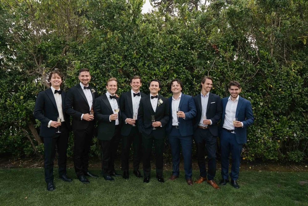 Groomsmen in dark suits and blue ties pose together in front of lush green foliage at an outdoor wedding venue.
