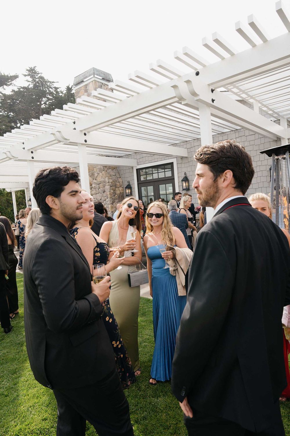 Outdoor wedding reception under a white pergola with guests mingling and socializing on a grassy lawn.