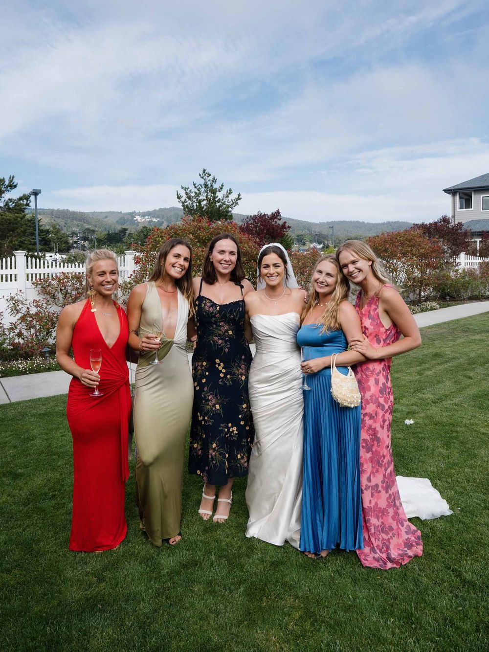 Group of friends in colorful formal dresses pose together on a grassy lawn with mountains in the background.