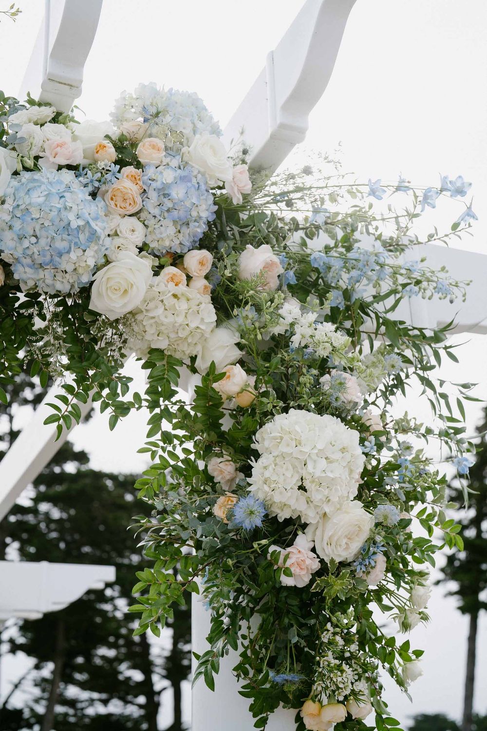 Elegant white and blue floral arrangements with roses and hydrangeas adorn a wedding arch with flowing fabric draping.