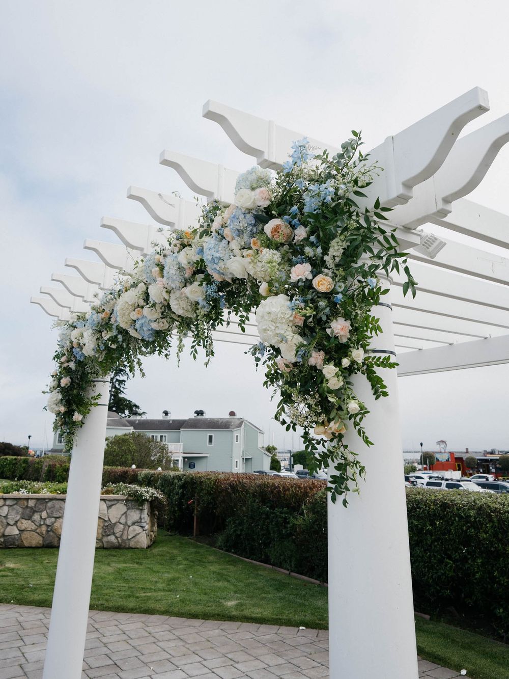 White wedding pergola decorated with elegant floral arrangements of white blooms and greenery against a cloudy sky.