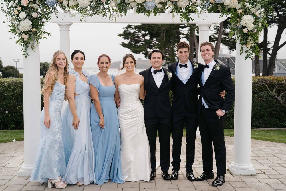 Wedding party in light blue bridesmaid dresses and black tuxedos posing outdoors.