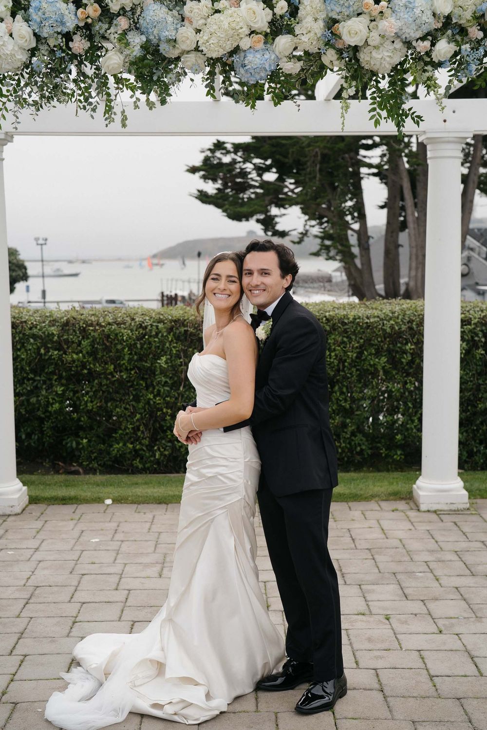 Newlyweds share a romantic moment under a white pergola decorated with floral arrangements and greenery.