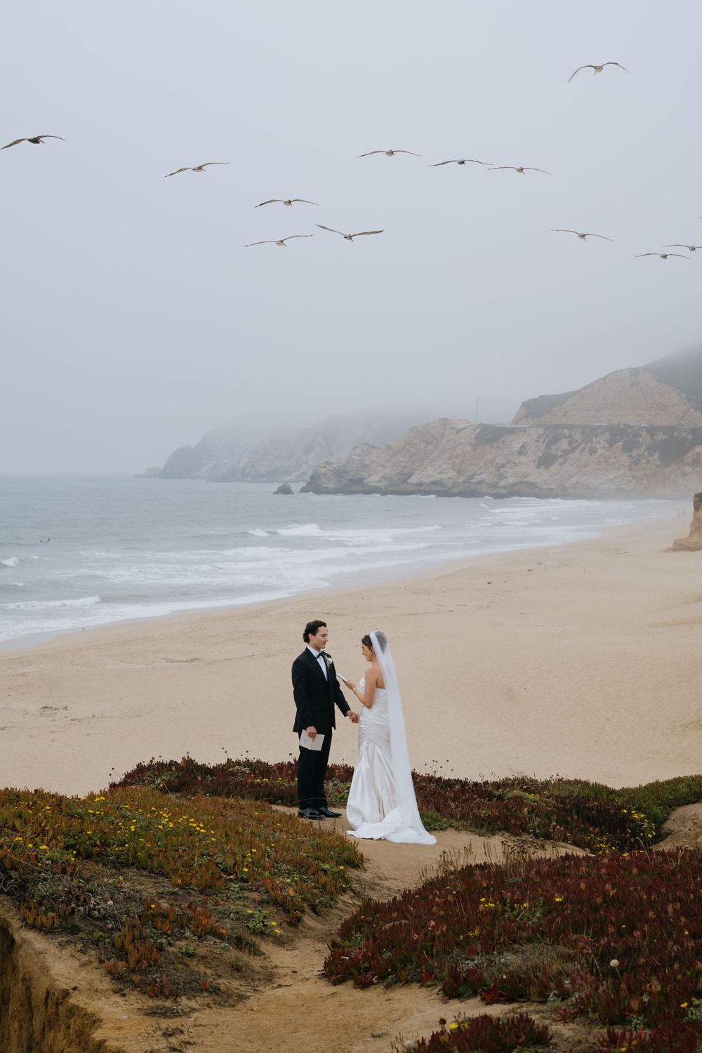 A couple in wedding attire stands on a misty beach with seagulls flying overhead and waves crashing on the shore.