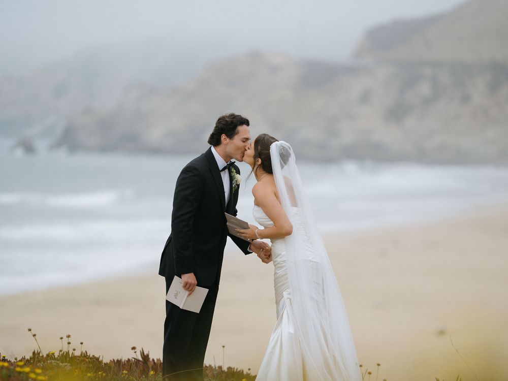 A romantic wedding couple embraces on a misty beach with cliffs in the background.