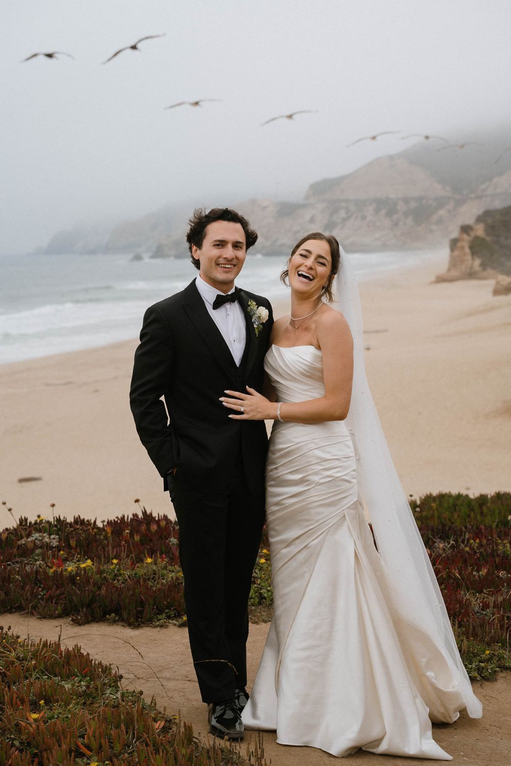 A couple shares joyful moments on a foggy beach with cliffs and seabirds in the background.