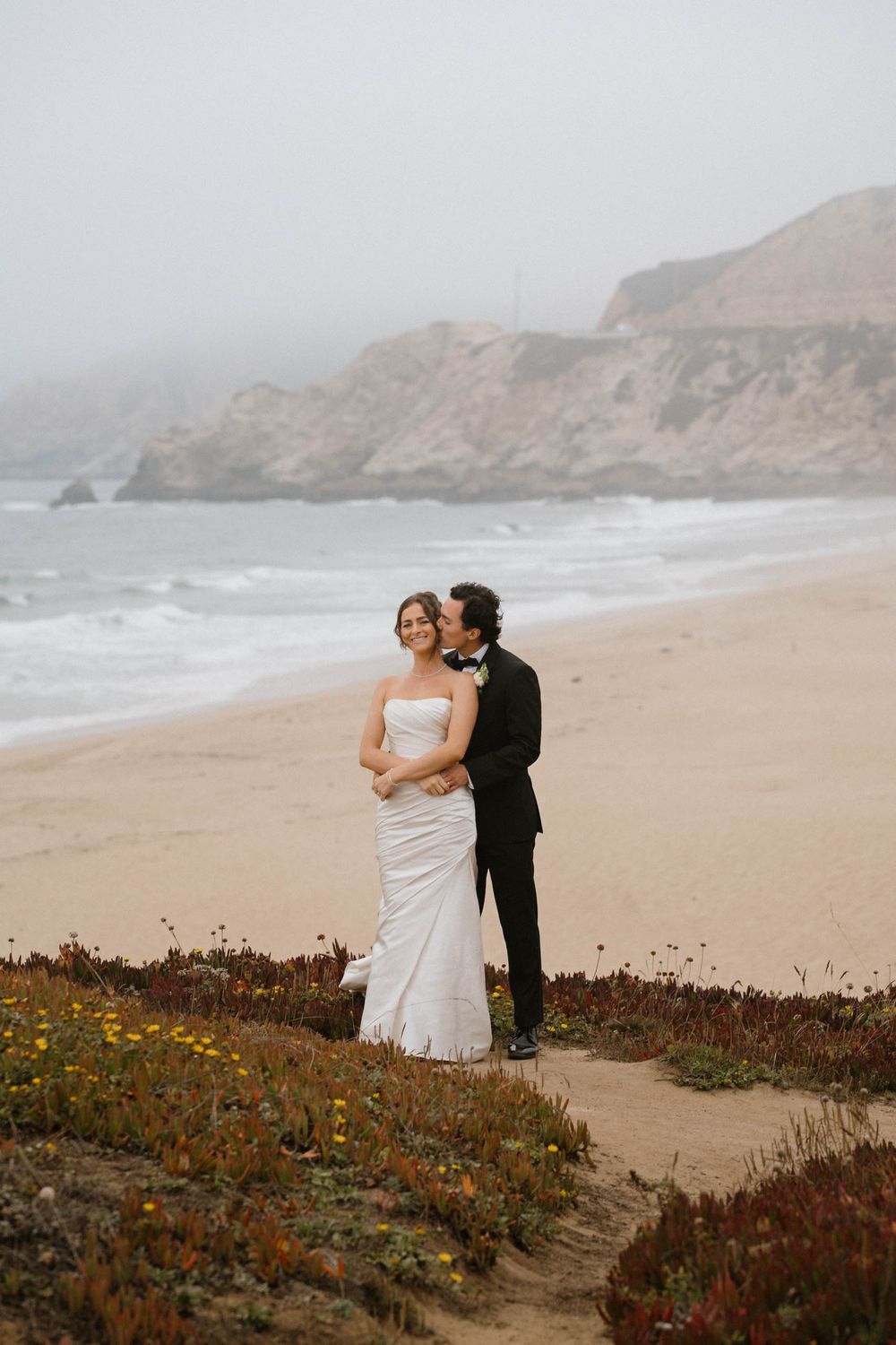 A couple in wedding attire embraces on a misty beach cliff with dramatic coastal scenery in the background.