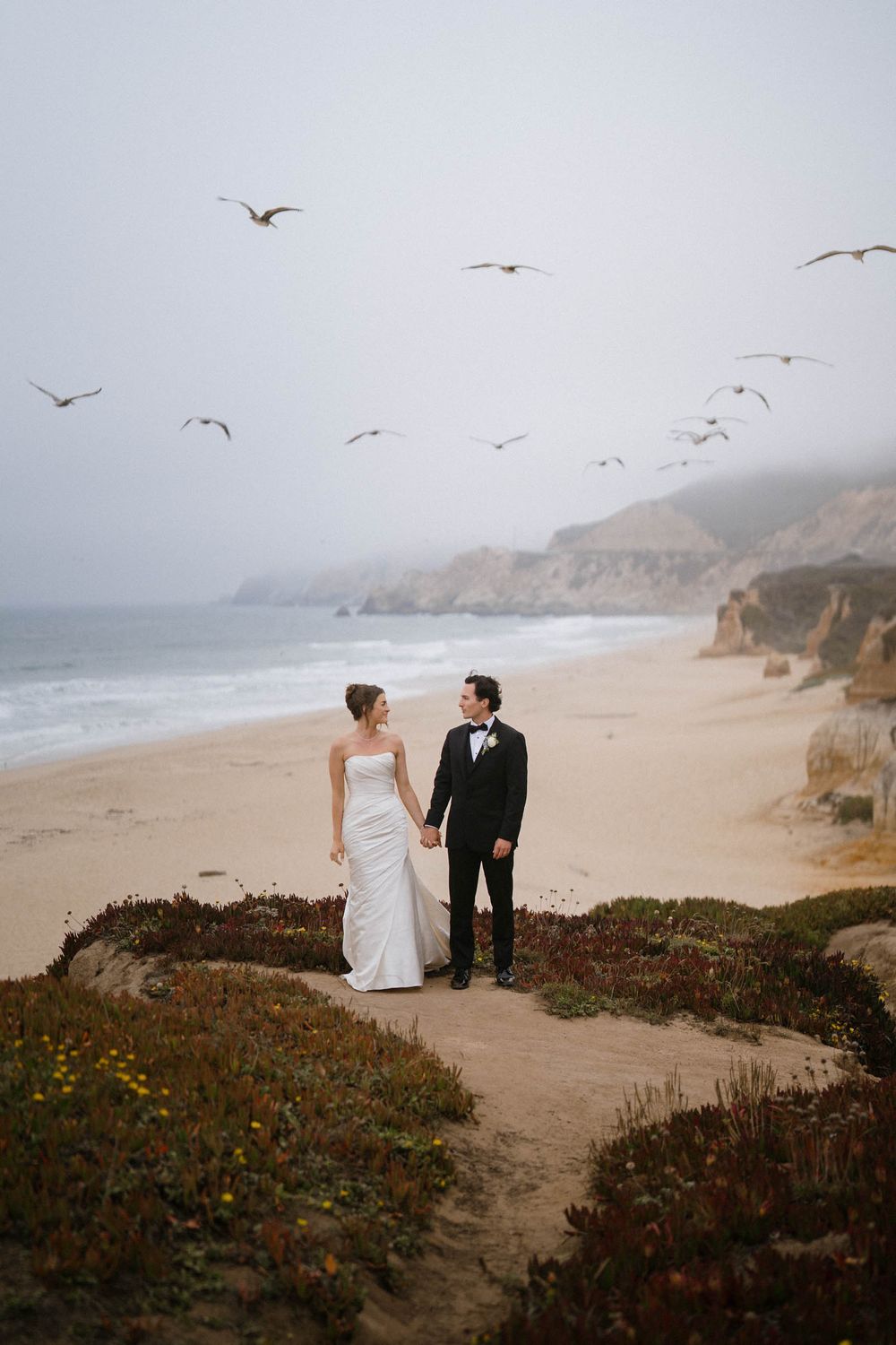 A couple in wedding attire walks along a misty coastal cliff path as seagulls soar overhead at sunset.