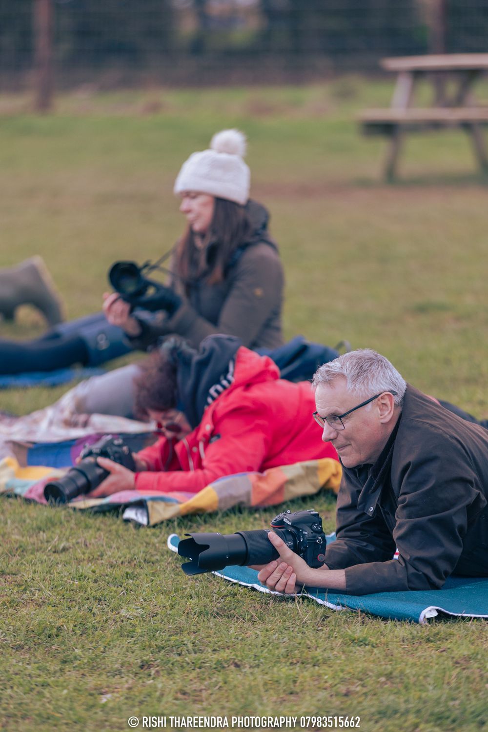 Photographers lying on ground mats with cameras during an outdoor photo session in a grassy field.