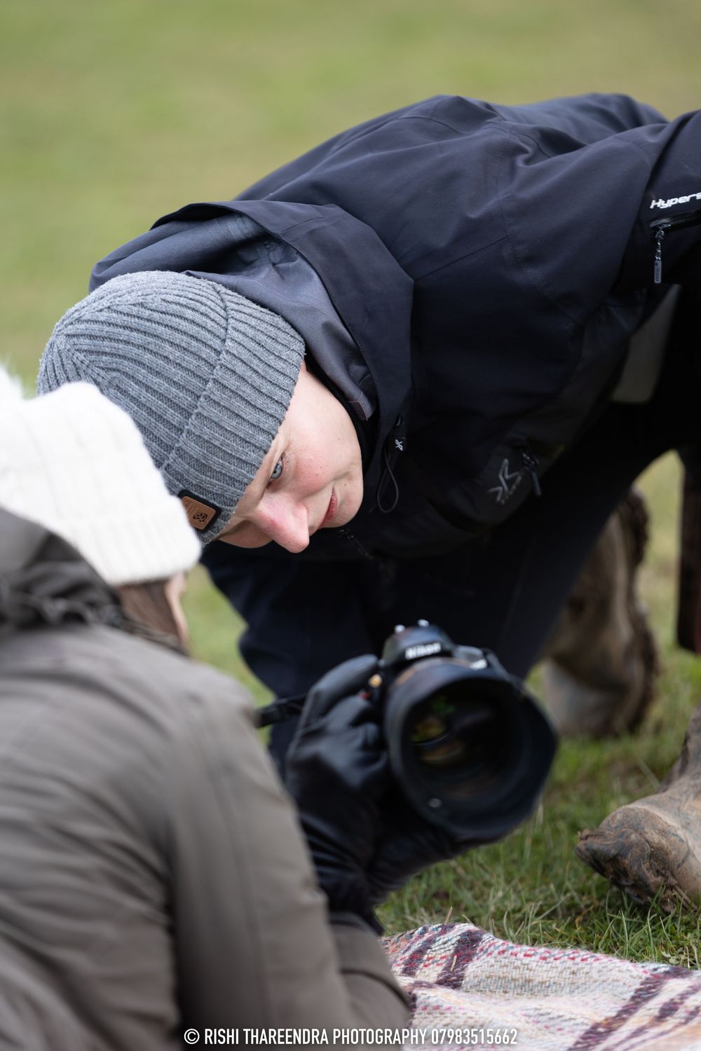 A person wearing a gray knit hat and black jacket lies on a blanket outdoors while petting a black dog.