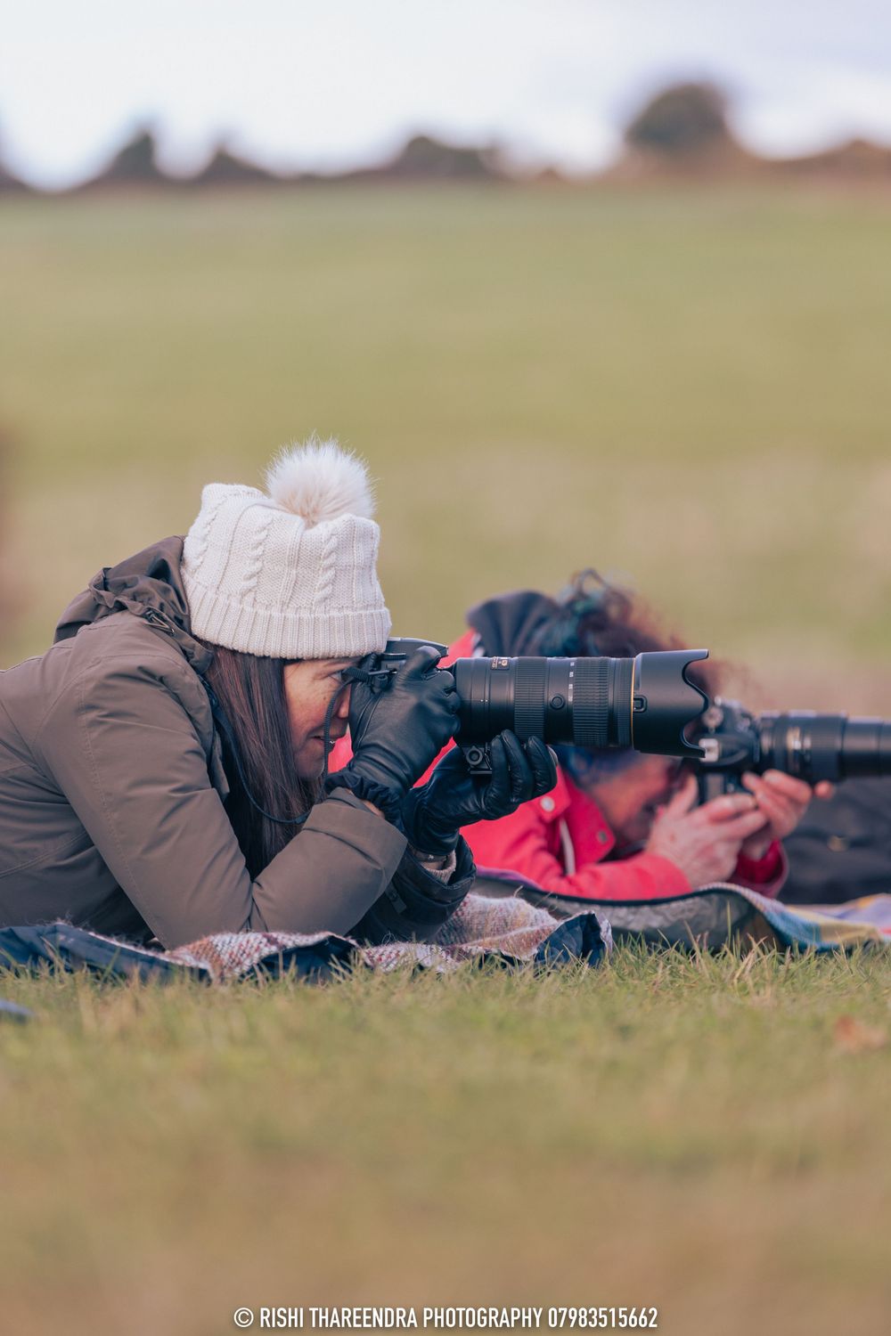 Wildlife photographer lying prone on grass with telephoto lens capturing nature shots in outdoor setting.
