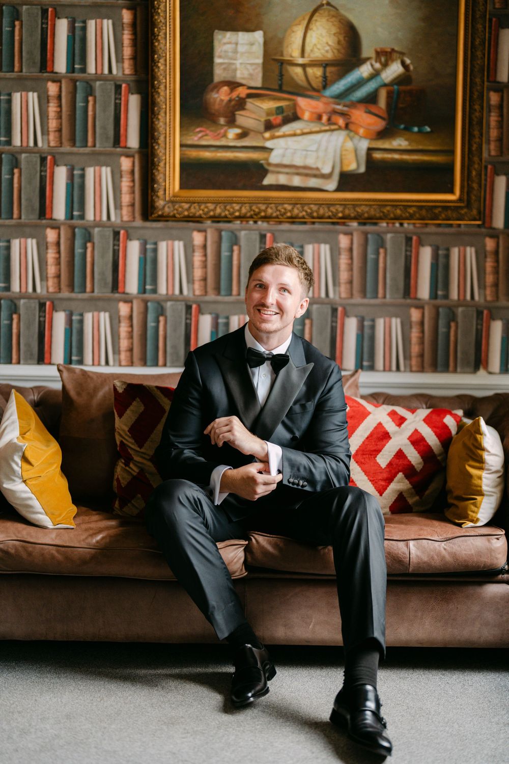 Formal portrait series of groom seated on brown leather couch with decorative pillows in library setting.