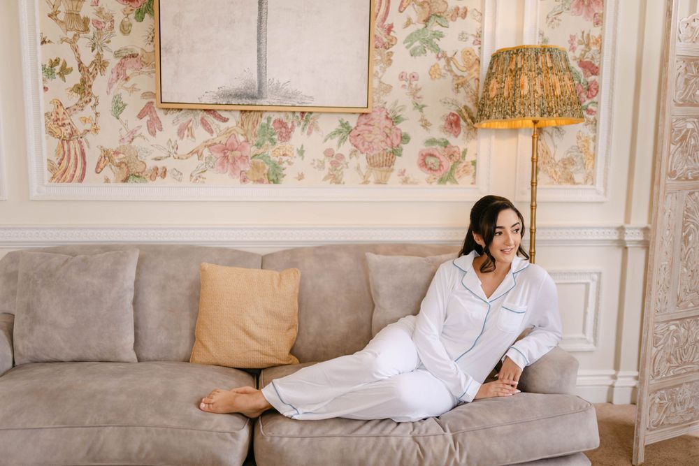 Bride in white robe relaxes on beige sofa against floral wallpaper in elegant room.