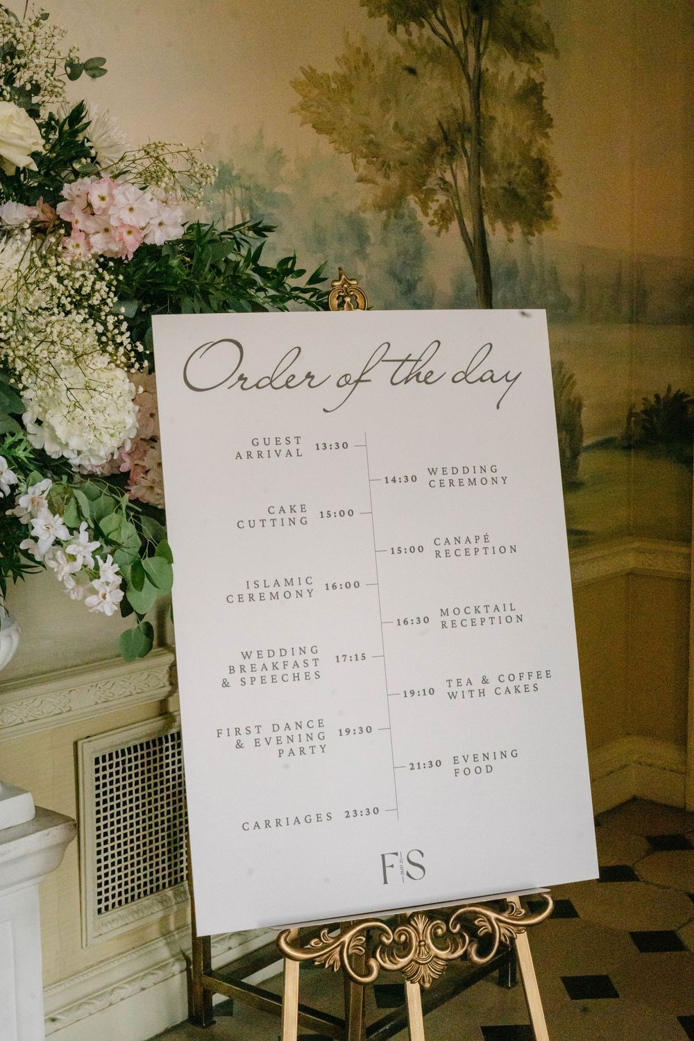 Wedding day timeline sign displayed on gold easel with floral arrangements and pastoral wallpaper.