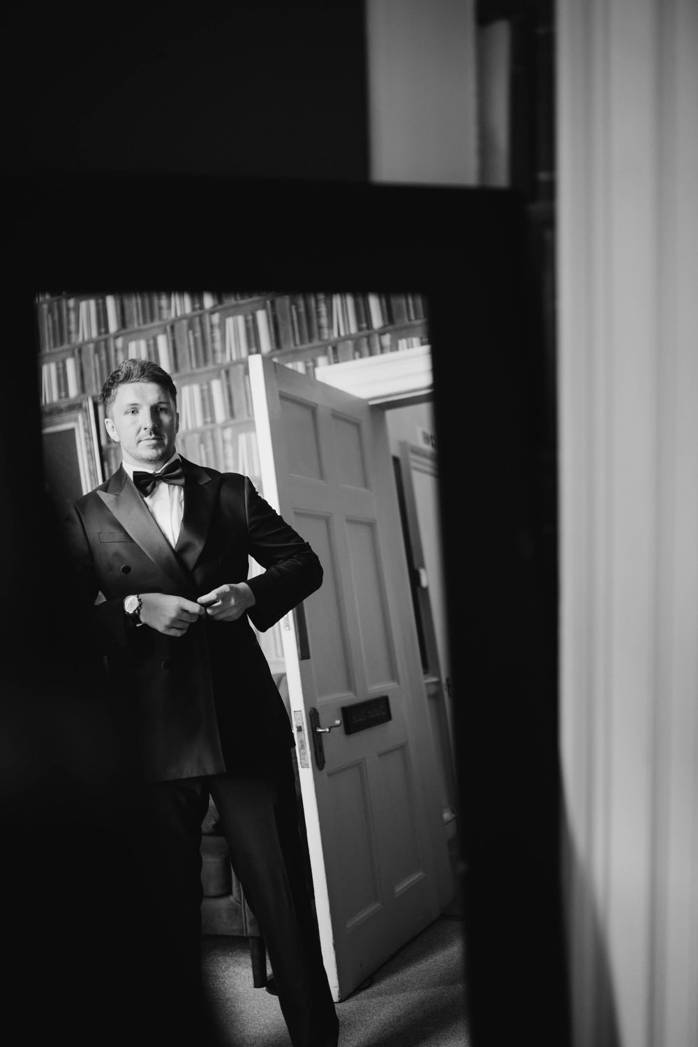 Black and white portrait of a groom in formal attire standing in a library setting.