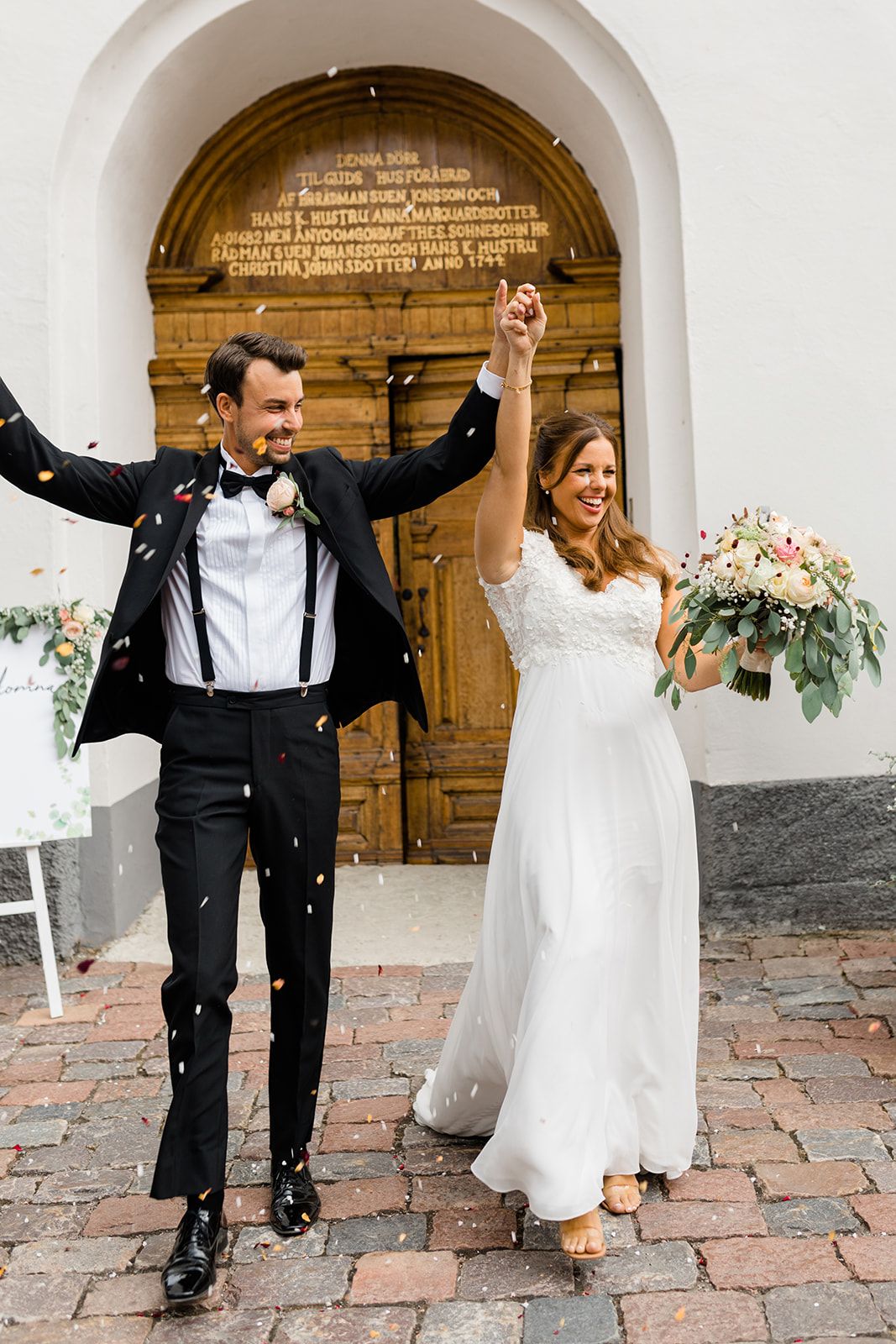 Newlyweds celebrate joyfully outside church entrance with confetti falling on brick pathway.