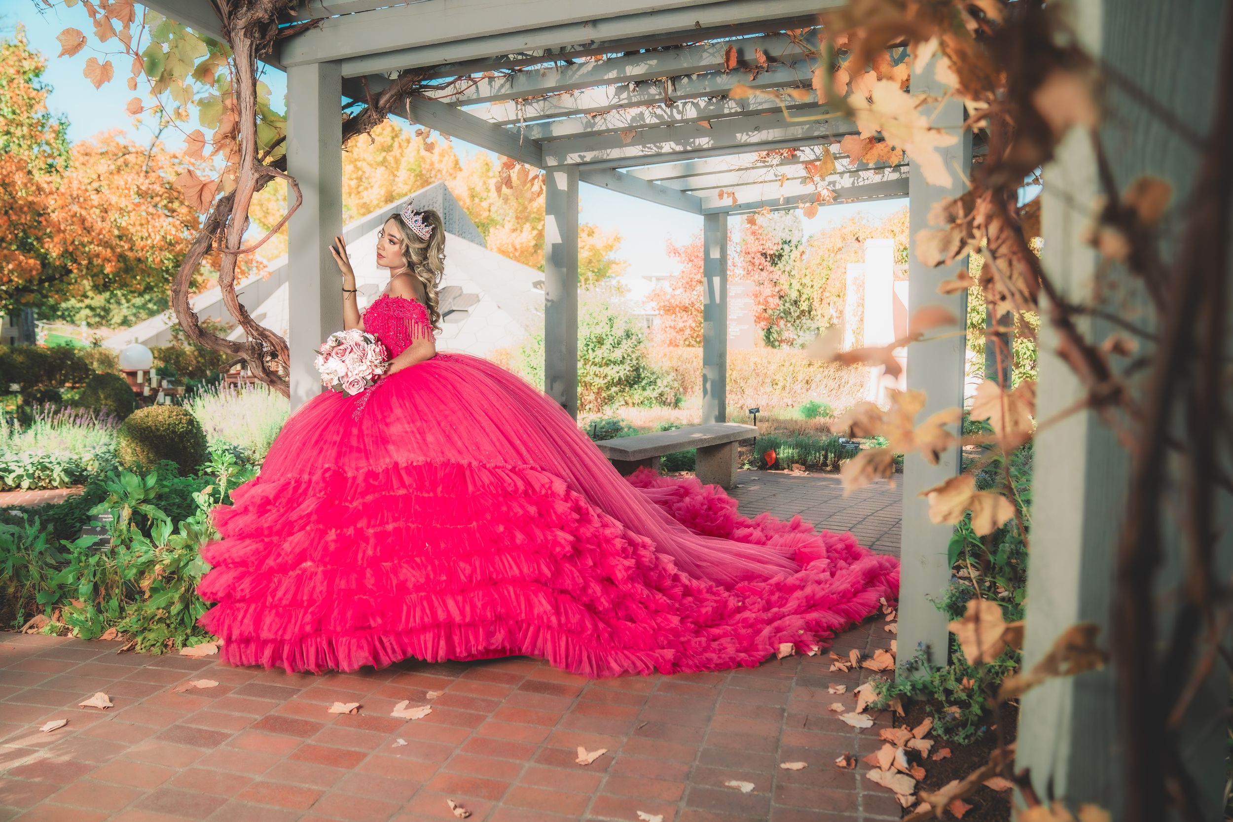 A bright pink tulle dress flows dramatically across a garden pathway beneath a rustic wooden pergola.