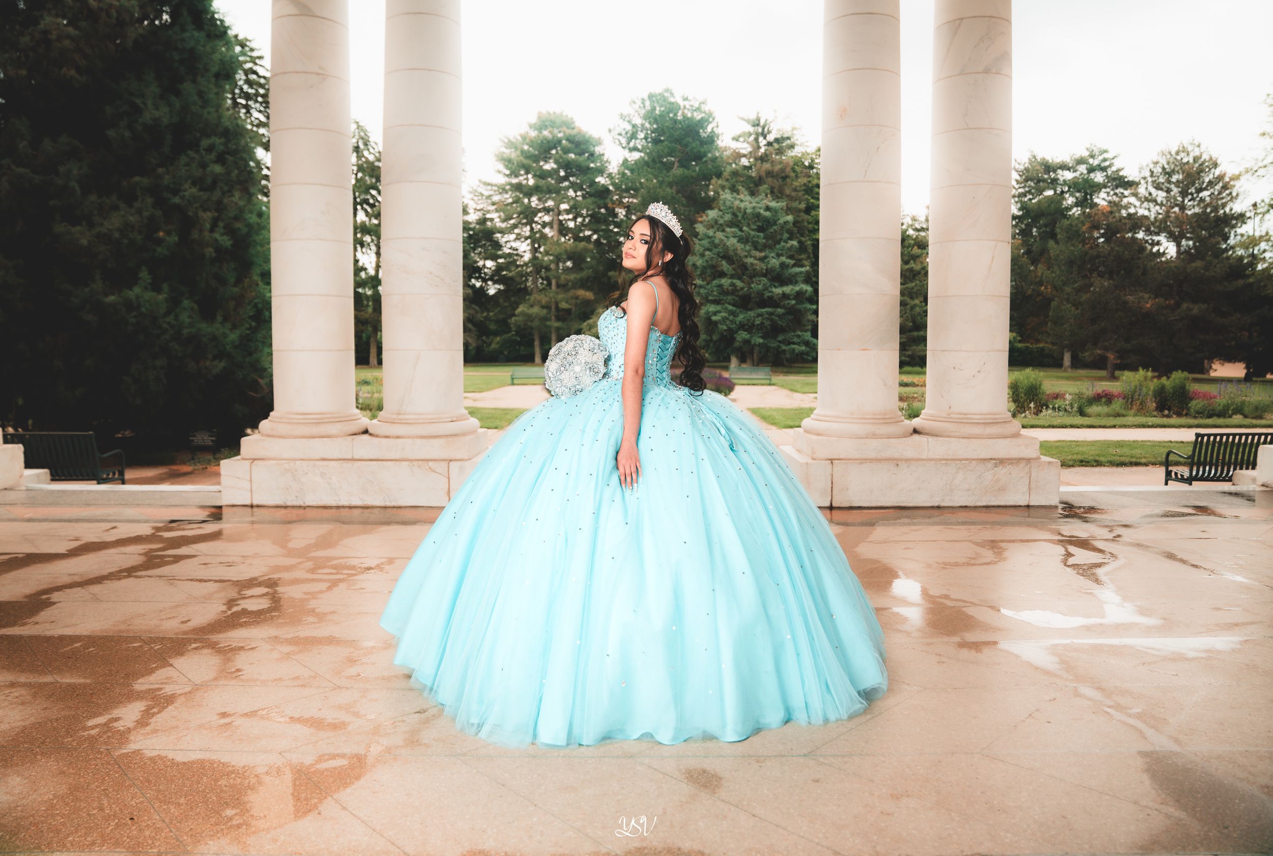 A person in a flowing turquoise quinceañera gown poses elegantly between white pillars at an outdoor venue.