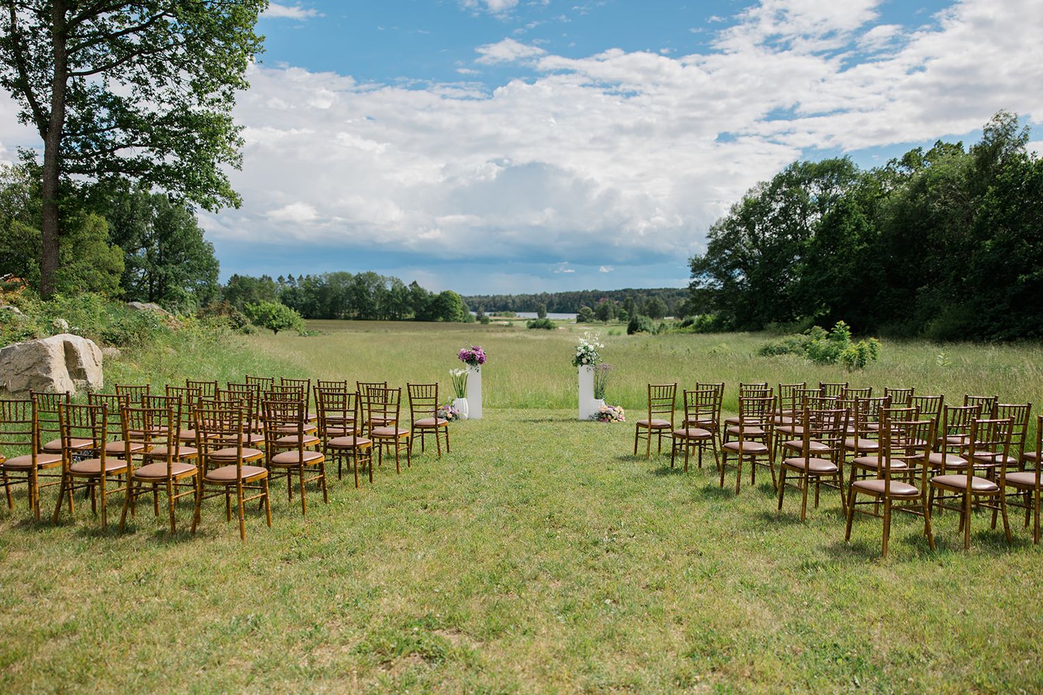 Wooden chairs arranged for an outdoor wedding ceremony in a scenic grassy field with mountain views and blue skies.