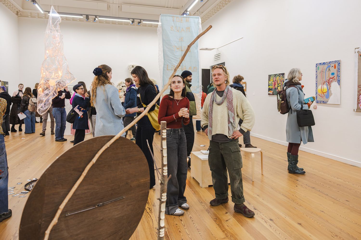 Gallery visitors observe artwork displayed in a modern white exhibition space with wooden floors.