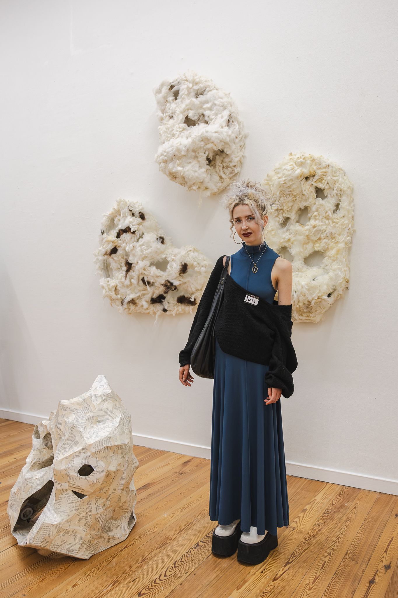 A person in a navy dress poses with large abstract white sculptural art pieces mounted on a gallery wall.