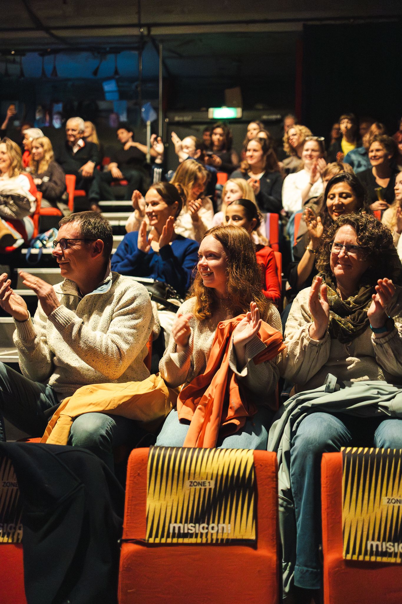 Audience members sit in yellow theater seats watching and reacting emotionally during a performance.