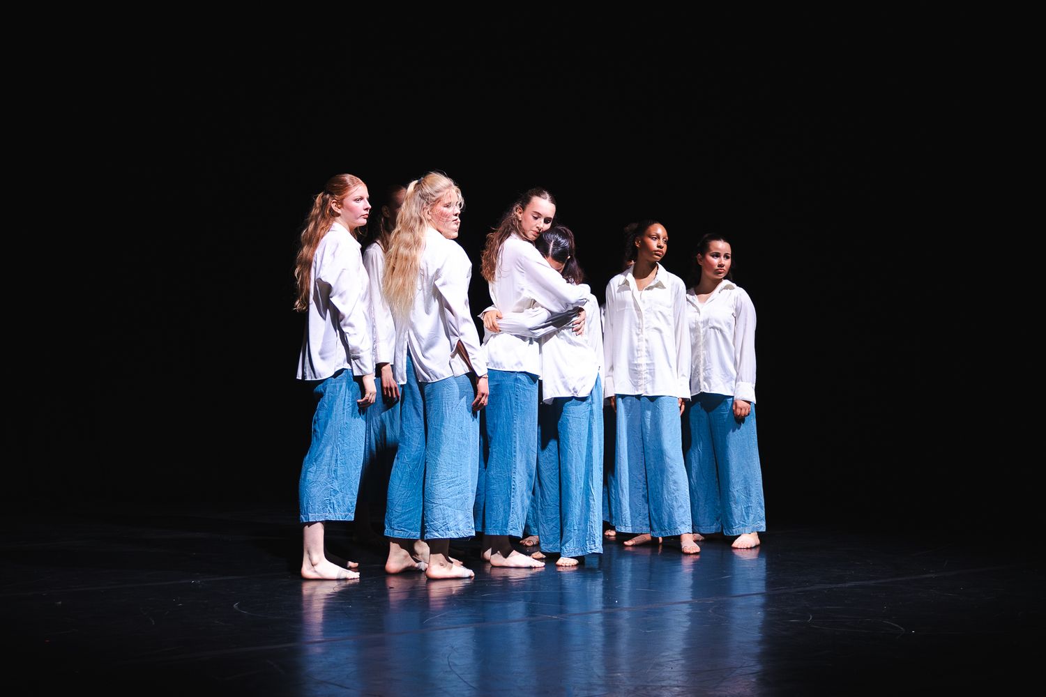Dancers in matching blue pants and white tops perform in formation on a dark stage during a recital.