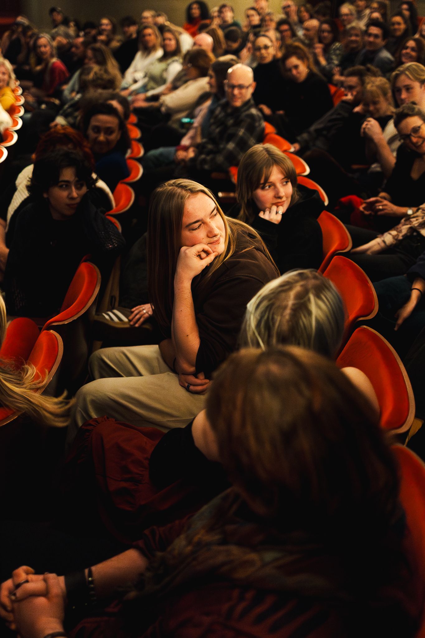 A crowded theater audience sits in red chairs during a performance, with people leaning and reacting emotionally.