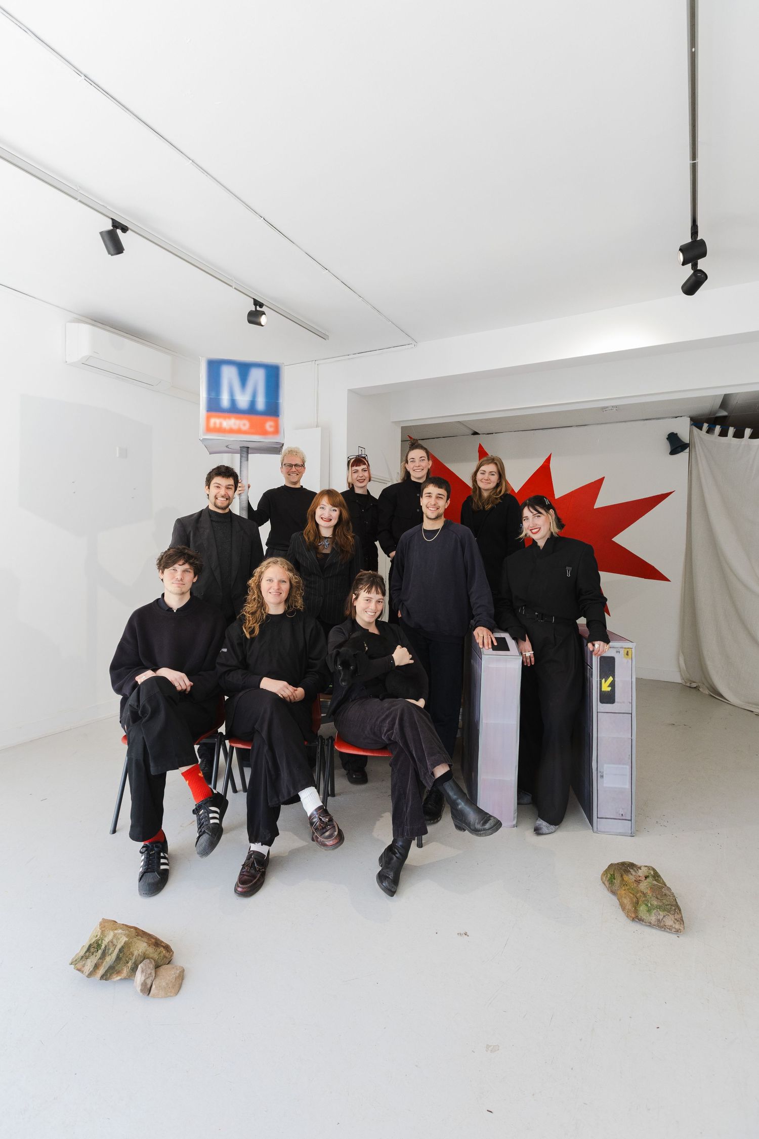 Group of people in black outfits posing in a white art gallery with a metro sign and red star sculpture.