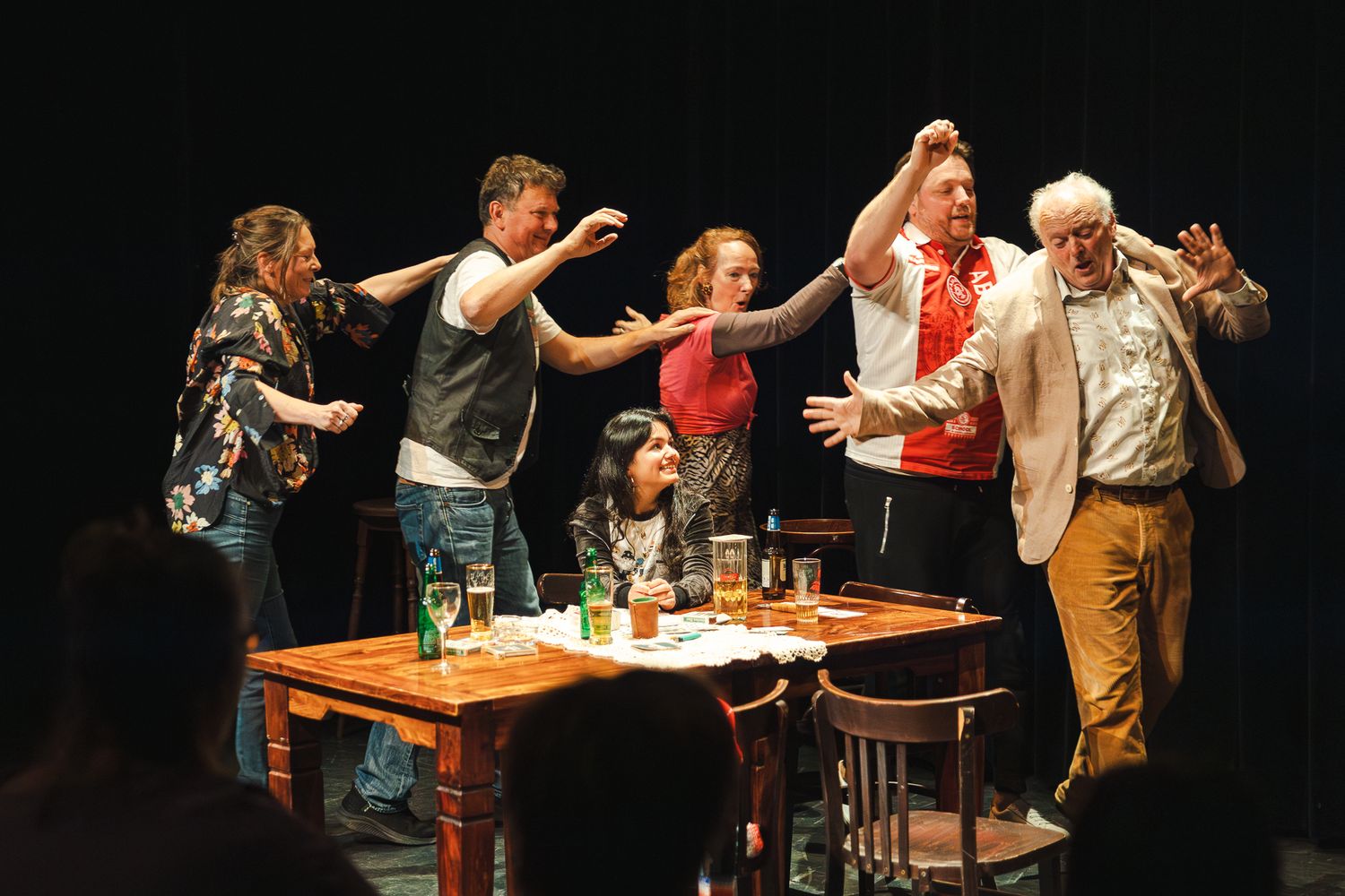 Performers on stage engage in an animated theatrical scene around a table with drinks during a dramatic performance.