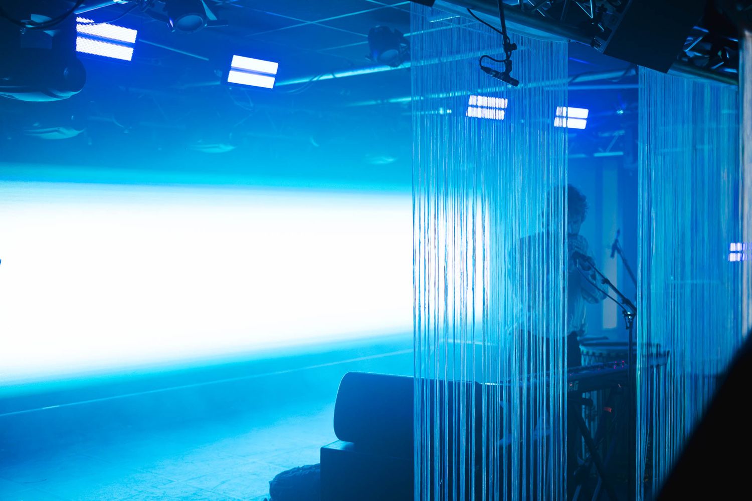 Blue-lit studio space with hanging string curtains, bright white screen, and professional lighting equipment.