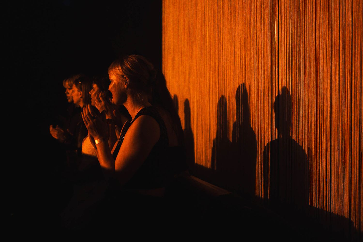 Women standing in dramatic orange light casting shadows on a curtained wall in a dark atmospheric setting.
