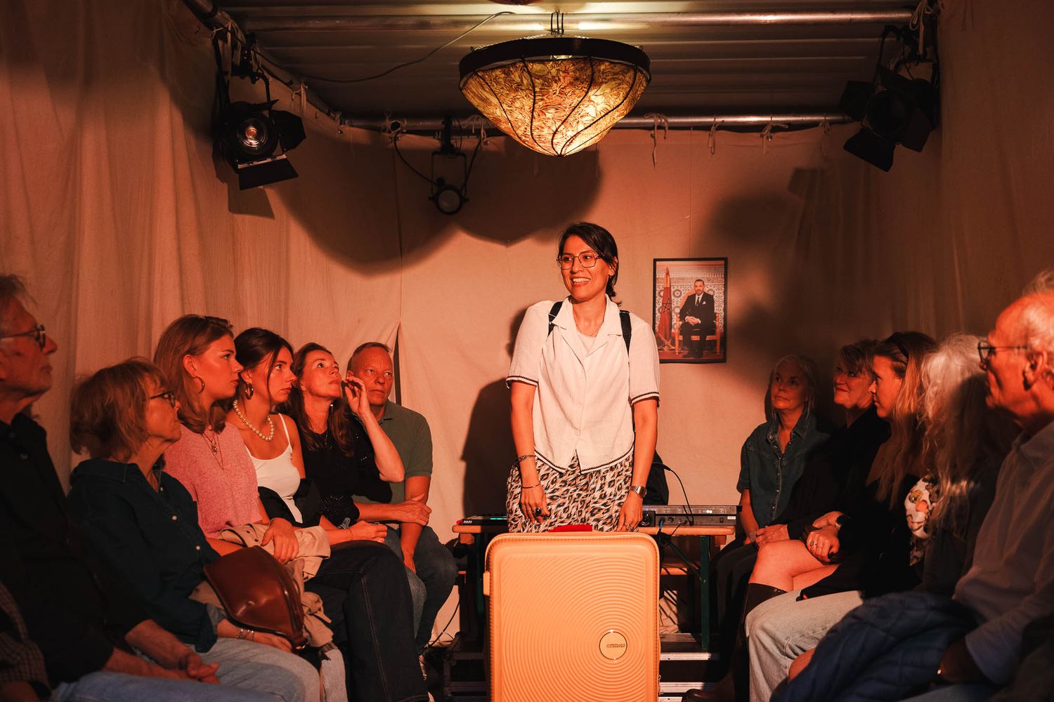 A woman stands confidently before a seated audience in a warmly lit intimate performance space.