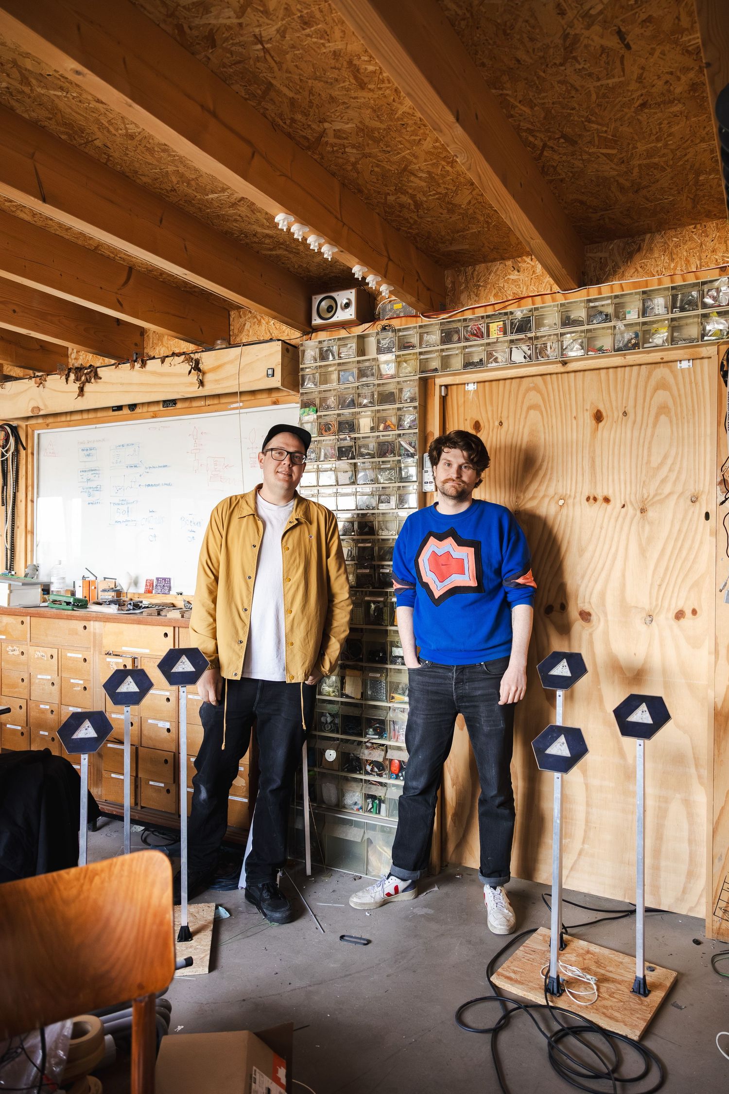 Two men standing in a wooden workshop studio with hexagonal speaker stands and a whiteboard in the background.