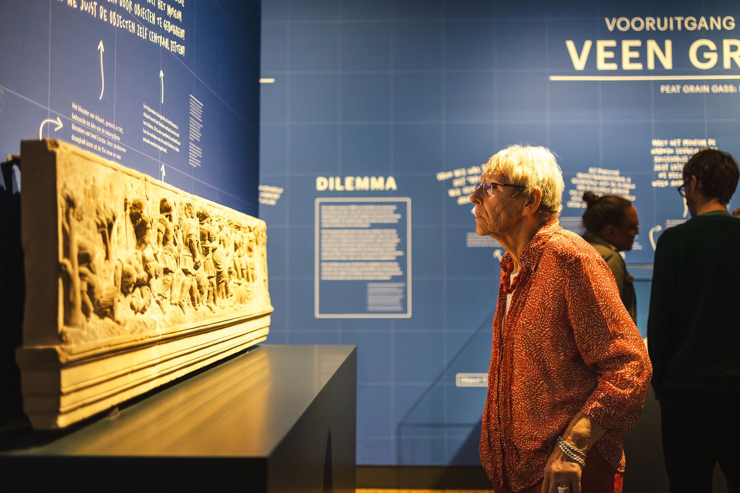Museum visitors examine a historical exhibit with illuminated displays and informational panels in a dimly lit gallery.