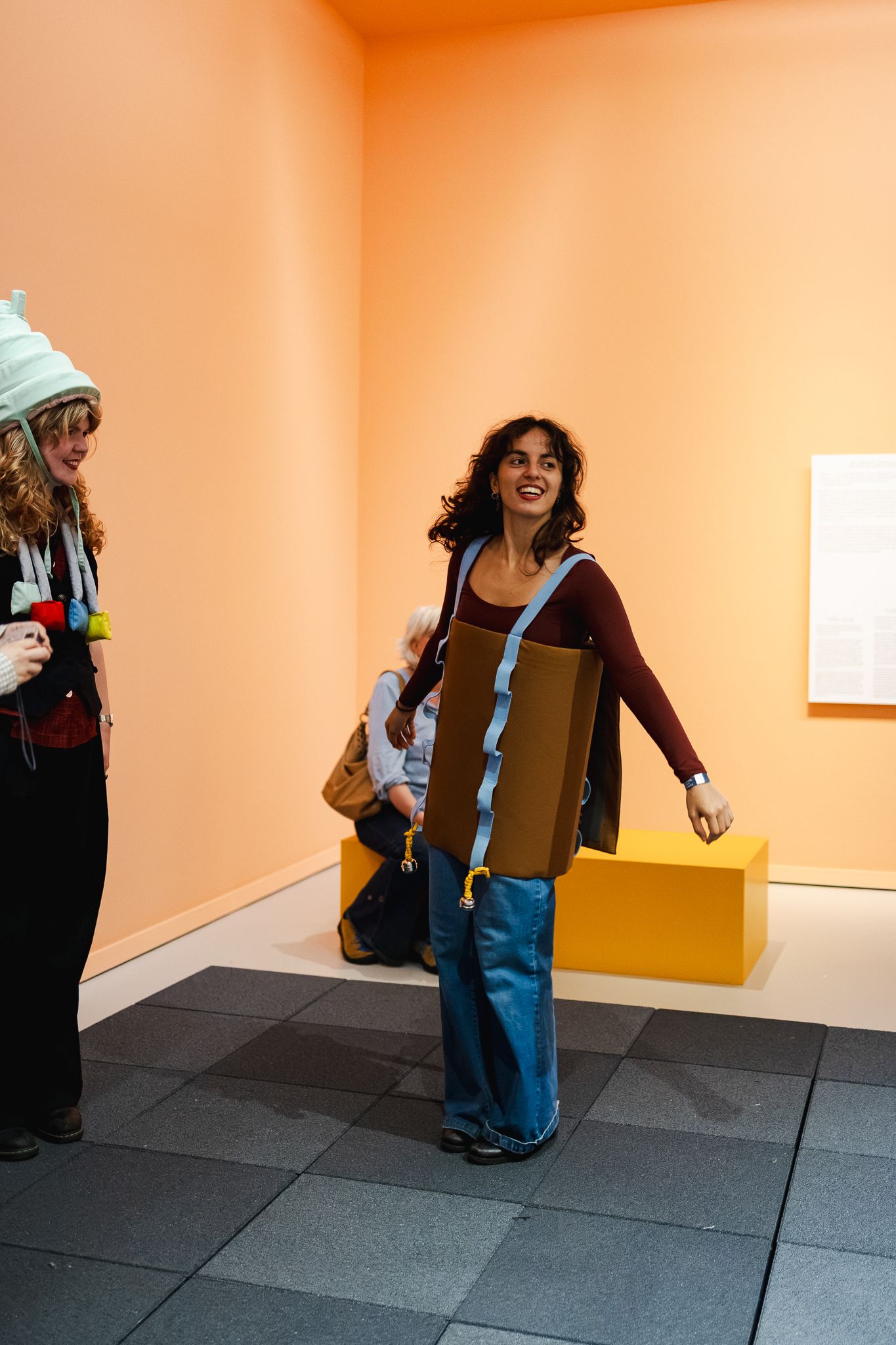 Woman in brown vest and wide-leg jeans smiles in an orange-walled art gallery with other visitors nearby.