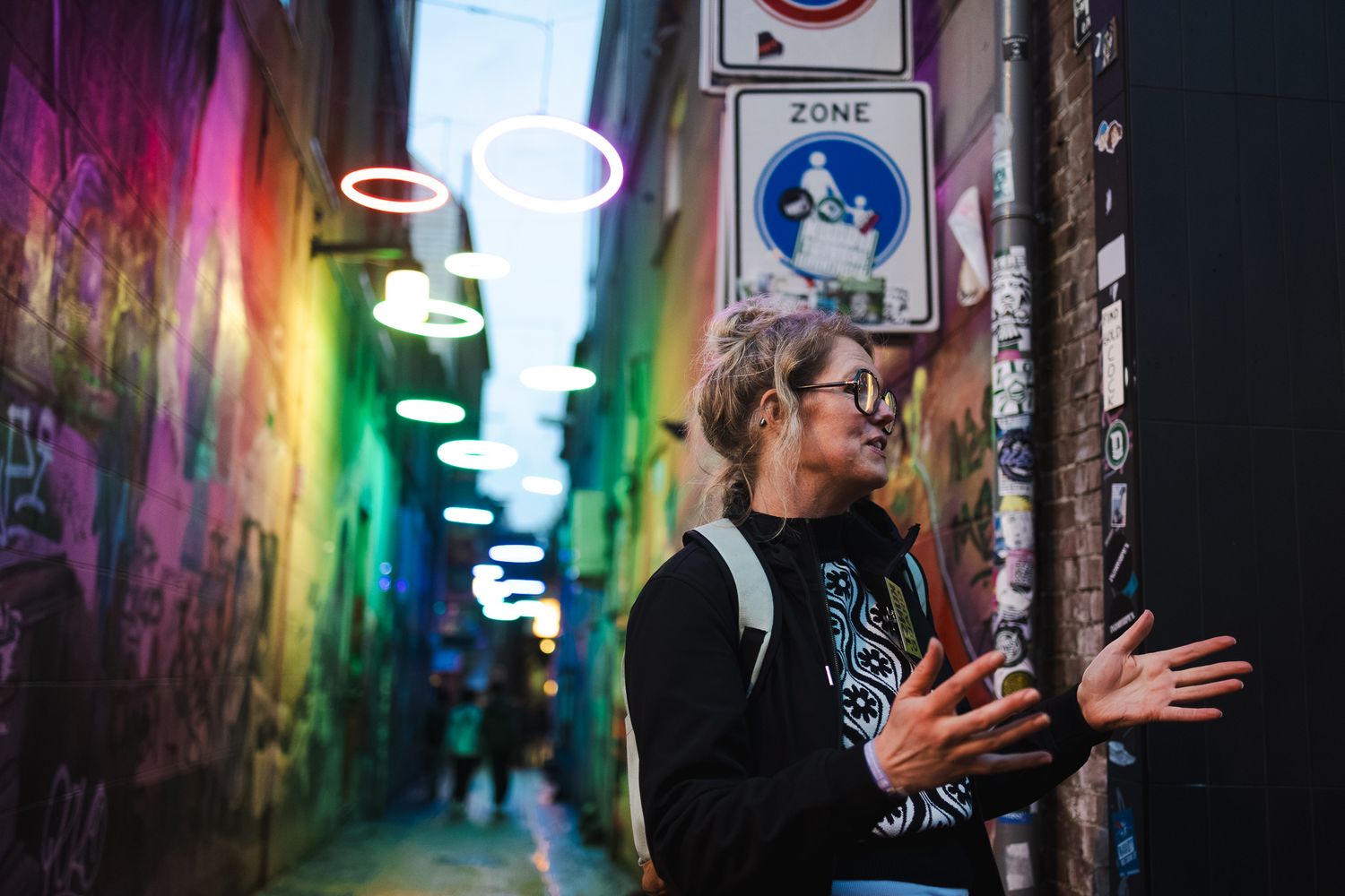 Woman with glasses explores a vibrant graffiti-filled alley at night, illuminated by colorful neon lights.